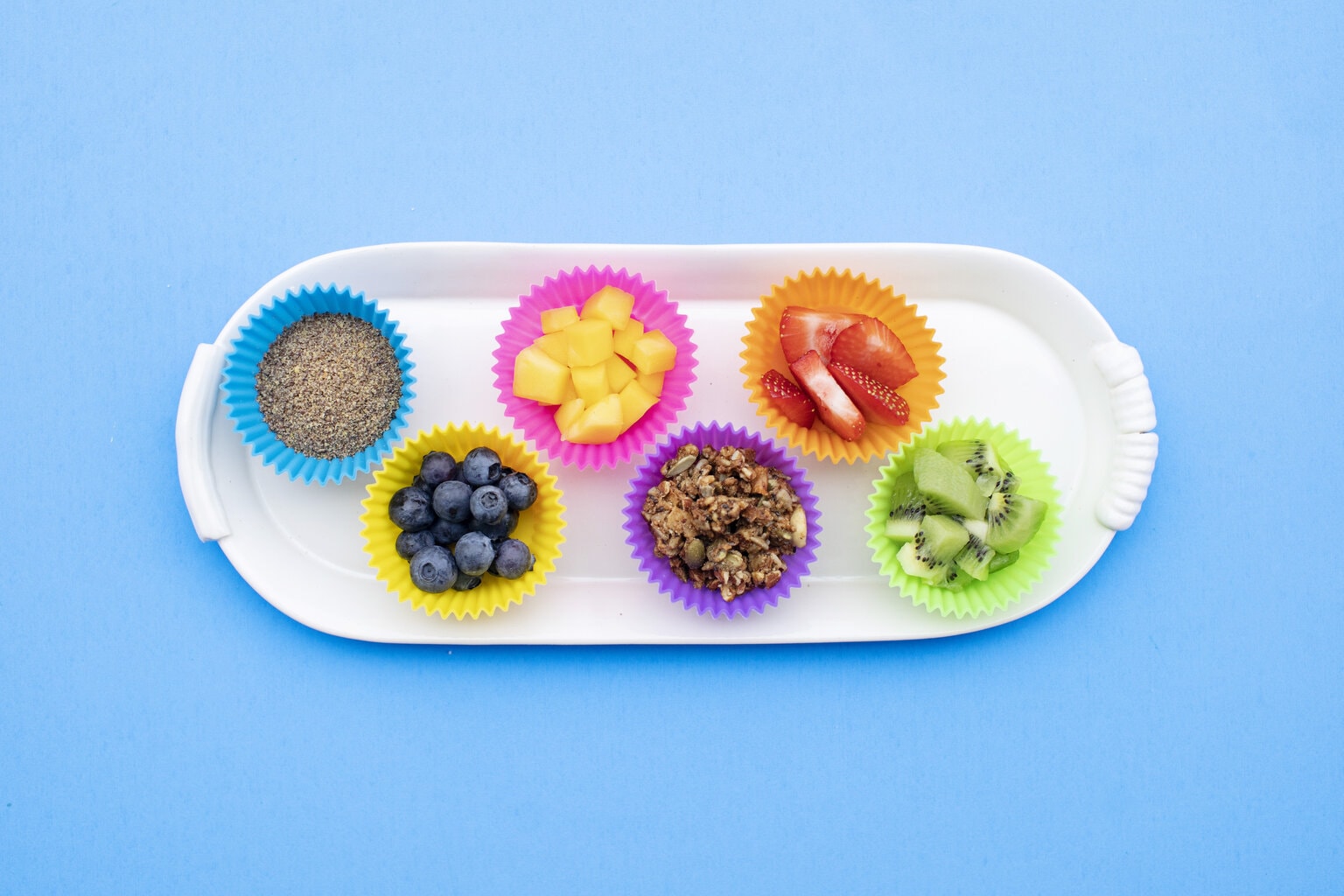 White tray with colorful fruit cups full of yogurt toppings against a blue background. 