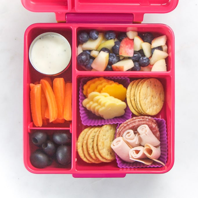 Pink school lunch box filled with healthy and easy school lunch ideas - cheese, crackers, fruit and carrots and ranch.