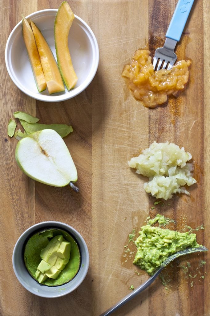 On a wooden cutting board we have one side slices of pear, cantaloupe and avocado and the other side is them smashed with a fork.