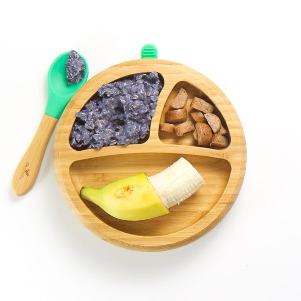 finger food breakfast on wooden plate with 3 sections - banana with end of peel still attached, blueberry oatmeal with spoon on the side of plate and chicken apple sausage cut into small chunks