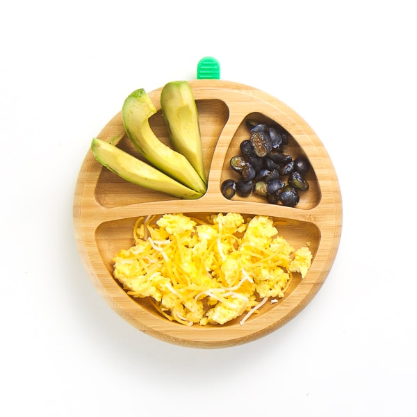 baby-led weaning breakfast on wooden plate with 3 sections - cheesy scrambled eggs, avocado wedges and cut blueberries