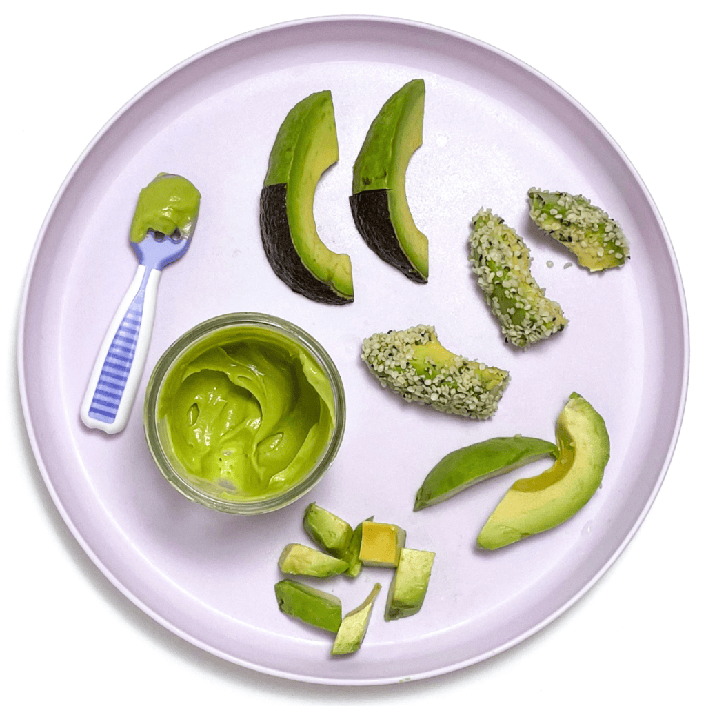 A purple baby plate with avocado made as a puree and as a finger food for baby-led weaning sitting on my white kitchen counter. 