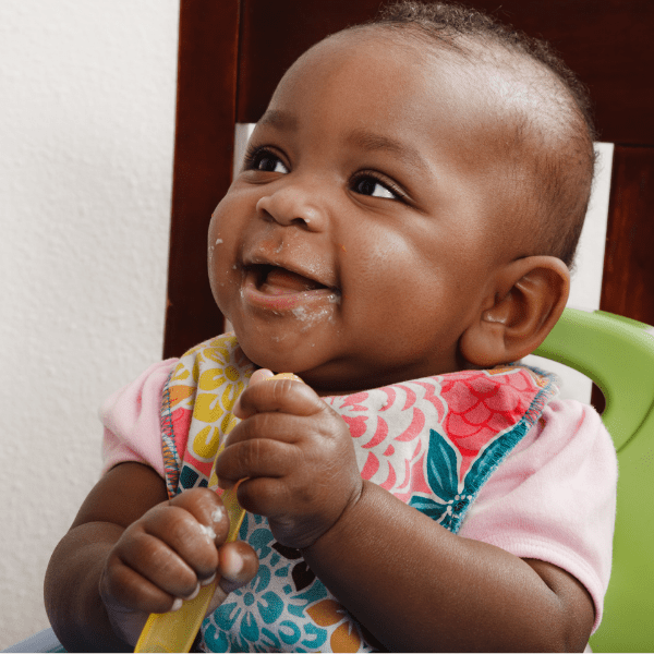 Baby sitting in a green booster seat is eating using a palmar grasp.