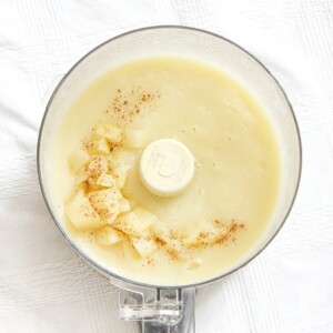 food processor sitting on a white tablecloth filled with pear baby food puree with chunks of pear and nutmeg sprinkled on top.