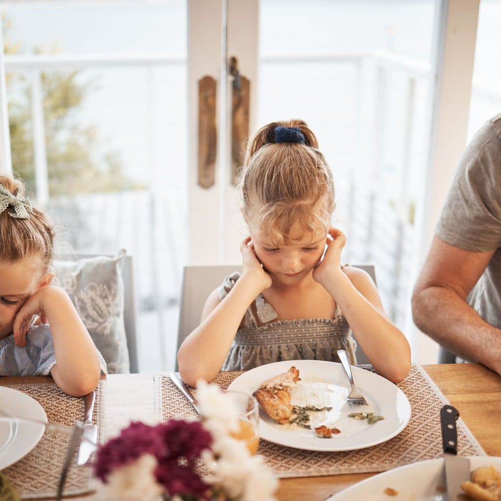 Image is of a girl at the dinner table staring at her plate of food but not eating. 