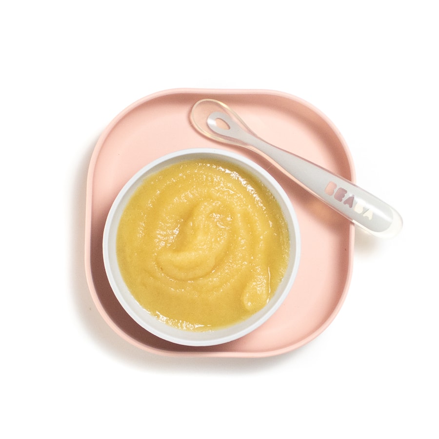 Pink baby play with a gray baby bowl on top with puréed apples and a gray spoon resting on the plate.