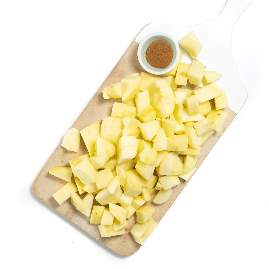 Wooden cutting board with chopped and peeled apples in a small bowl of cinnamon.