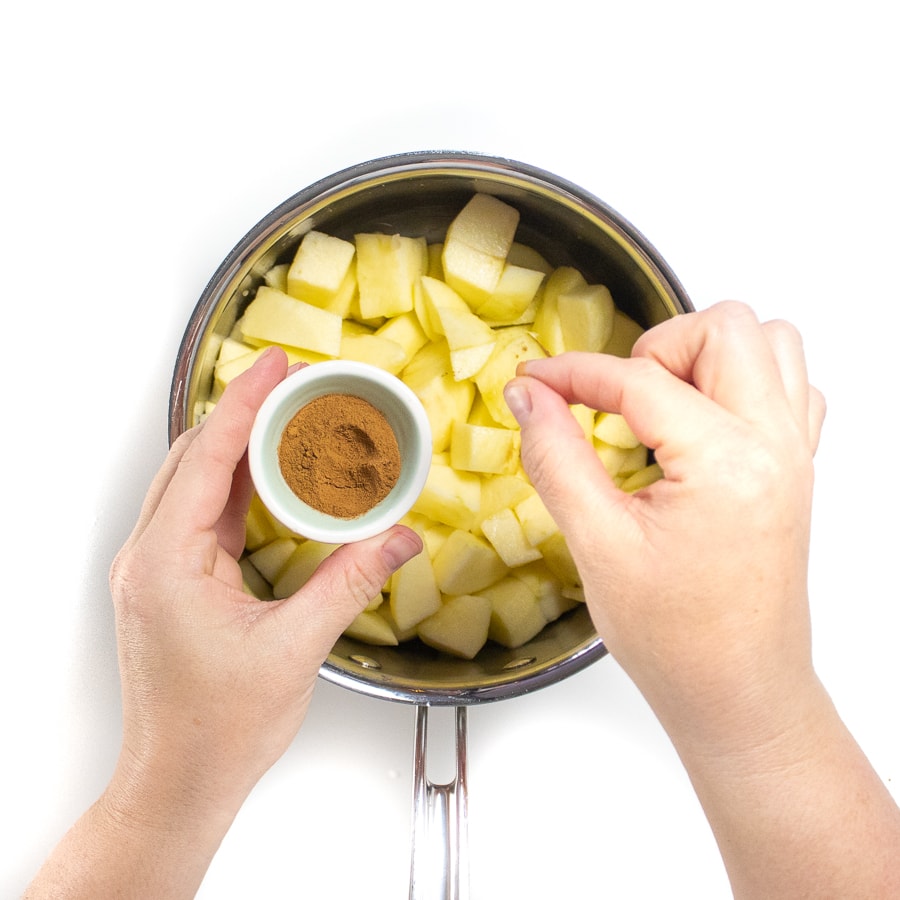 Silver sauce pan with chopped apples and hands reaching over with a pinch of cinnamon.