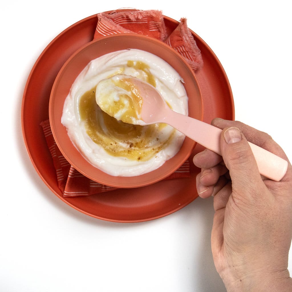 A hand holding a pink spoon full of apple yogurt over a bowl of apple yogurt.