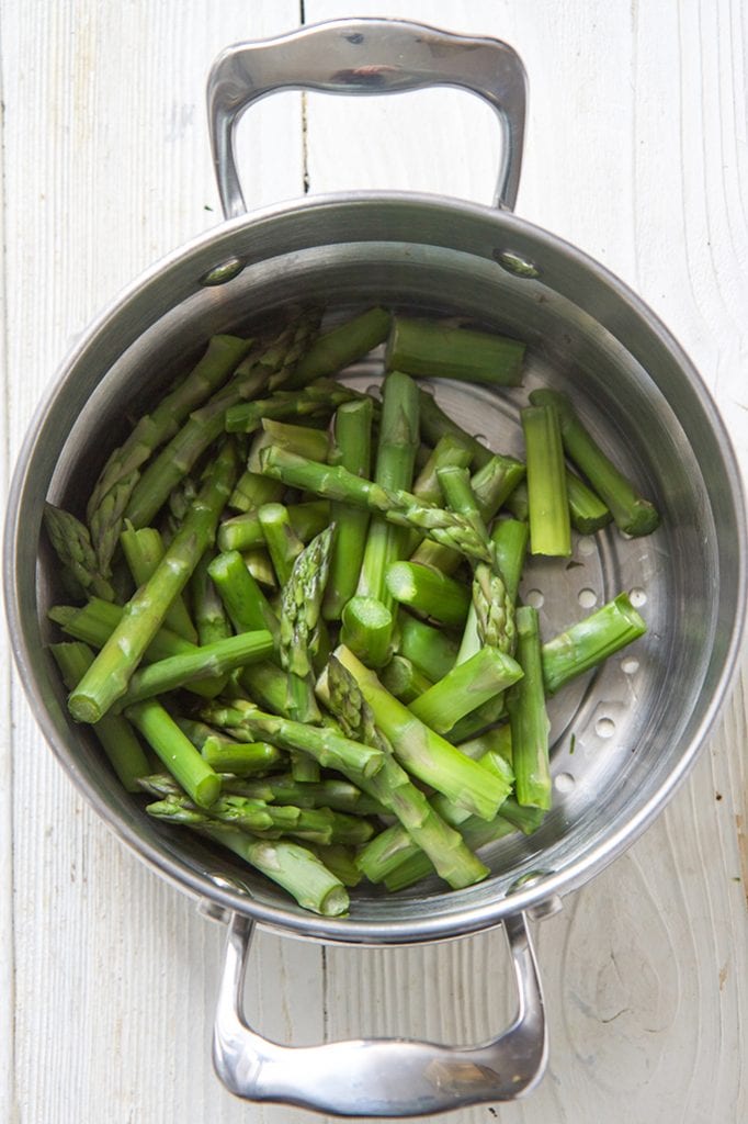 silver steamer basket filled with cut pieces of asparagus, sitting on a white wooden board