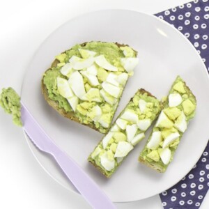 A white round plate with a cut up piece of avocado toast and egg sitting on top.