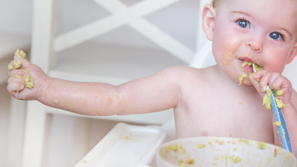 Baby sitting in a white high chair self feeding a soft puree. 