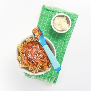 small white bowl filled with baby led weaning pasta with Bolognese sauce on top. Bowl has blue fork with pasta sitting on top of bowl.