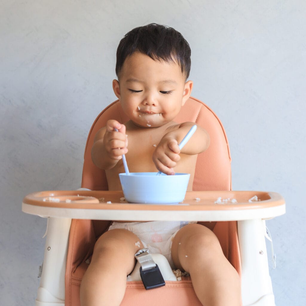 Small baby sitting in a high chair eating with two spoons.