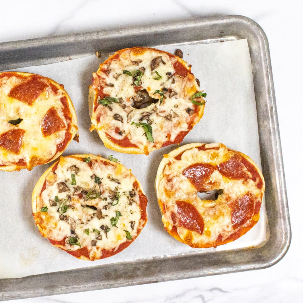 Four bagel pizzas on a baking sheet on a marble countertop with different toppings.