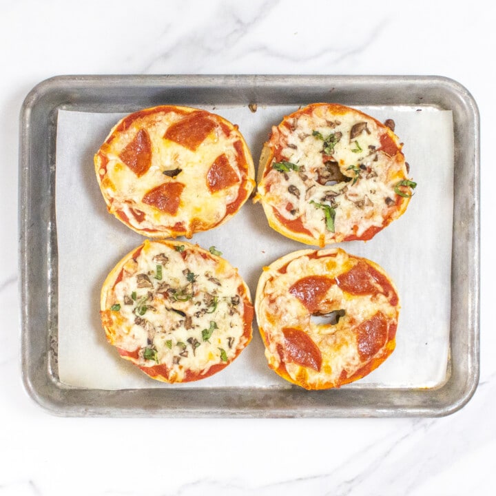 Four bagel pizzas on a baking sheet on a marble countertop with different toppings.