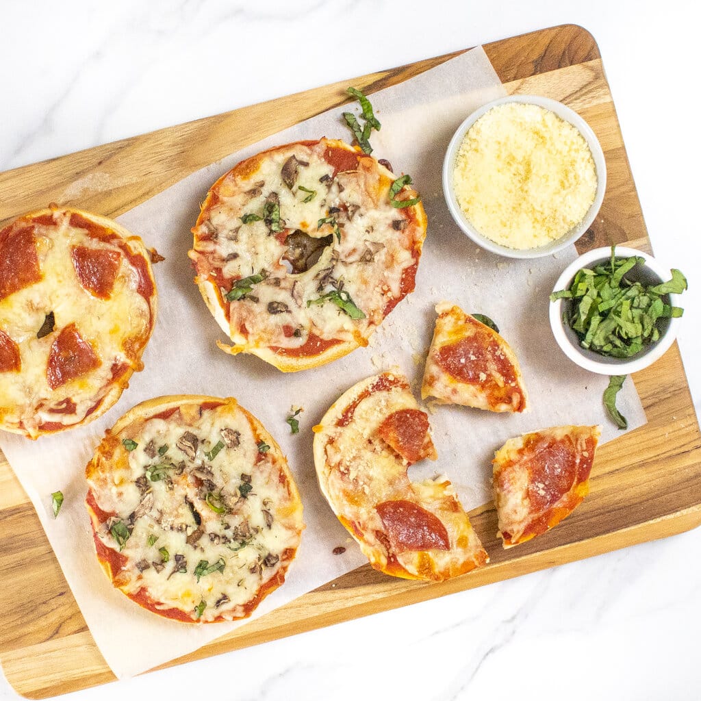 Bagel pizzas on a cutting board with Parmesan and basil getting ready to be cut for a kids meal.