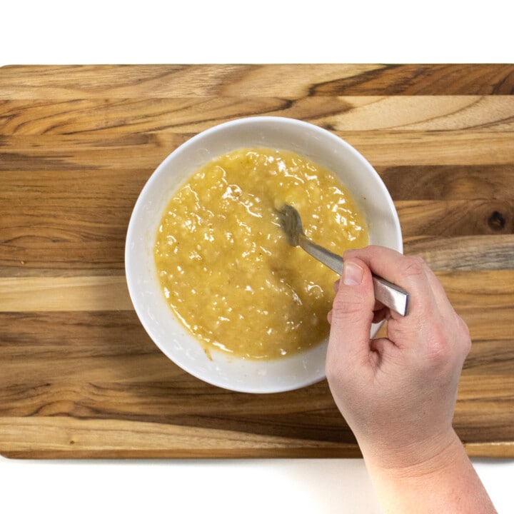 A hand mashing banana, maple syrup and nutmeg to mix into yogurt.