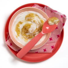 A pink kids bowl and plate with a pink napkin full of banana yogurt with a spoon resting on top.