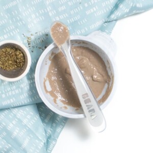 Gray baby bowl with baby spoon resting on top with a teal napkin on table.