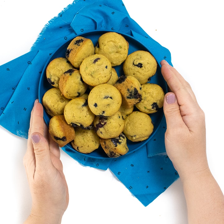 Hands holding a blue kids plate pilled high with blueberry muffins. 