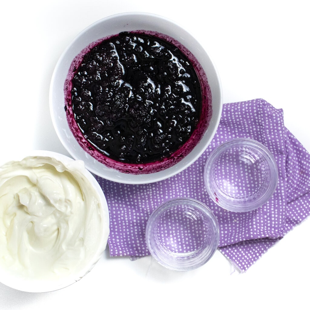 A bowl of blueberry compote and yogurt being put into small jars on a purple napkin against a white background.