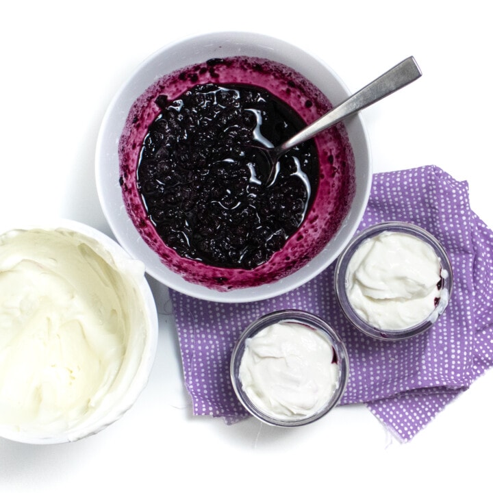 A bowl of blueberry compote and yogurt being put into small jars on a purple napkin against a white background.