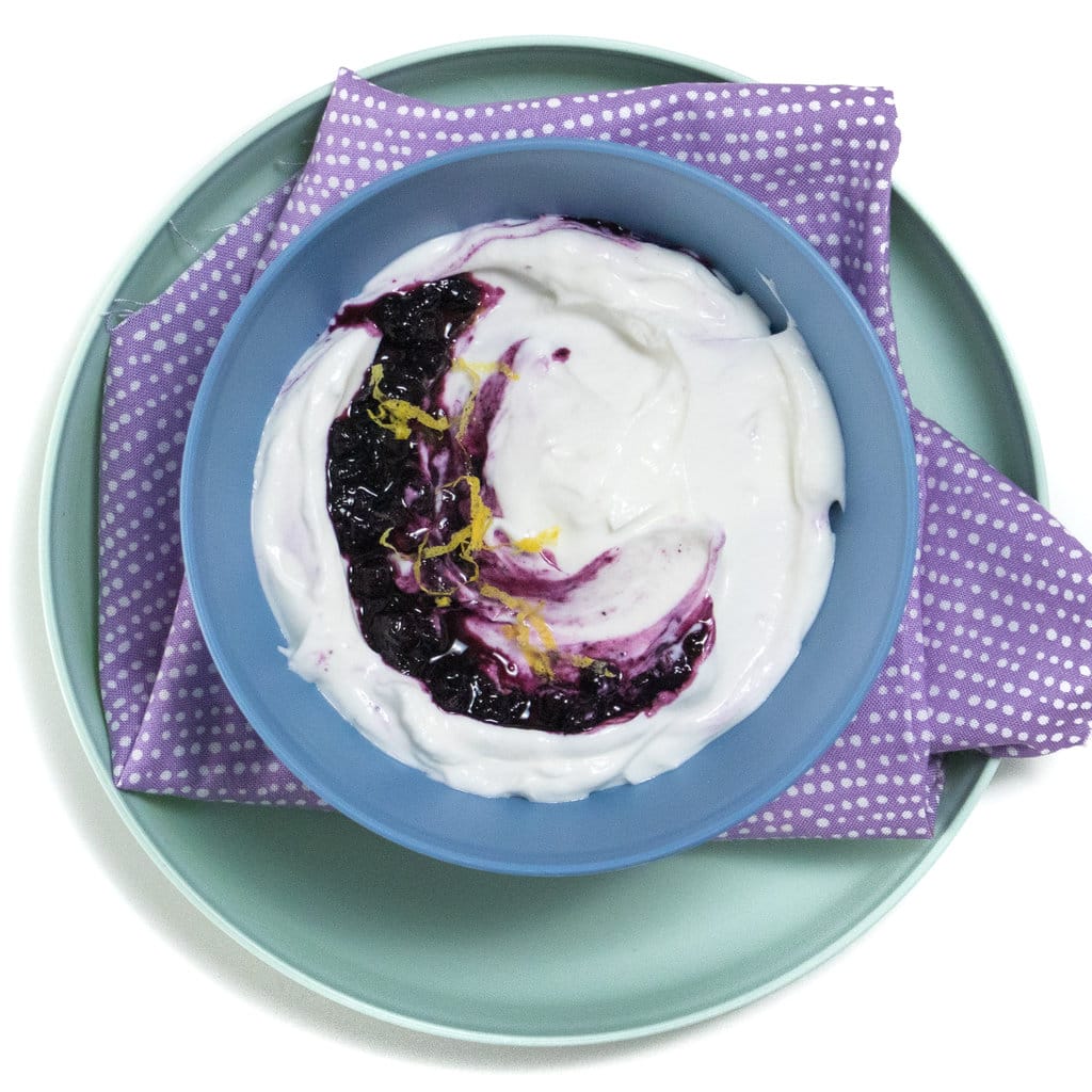 Blue kids bowl with a green kids played against a white background with blueberry yogurt with a sprinkle of lemon zest.