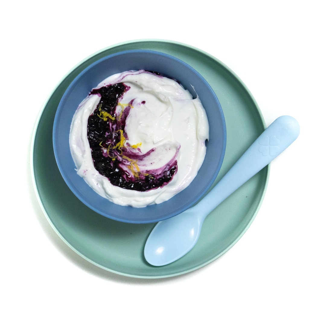 A white background with a teal kids plate with a blue kids bowl full of blueberry yogurt.