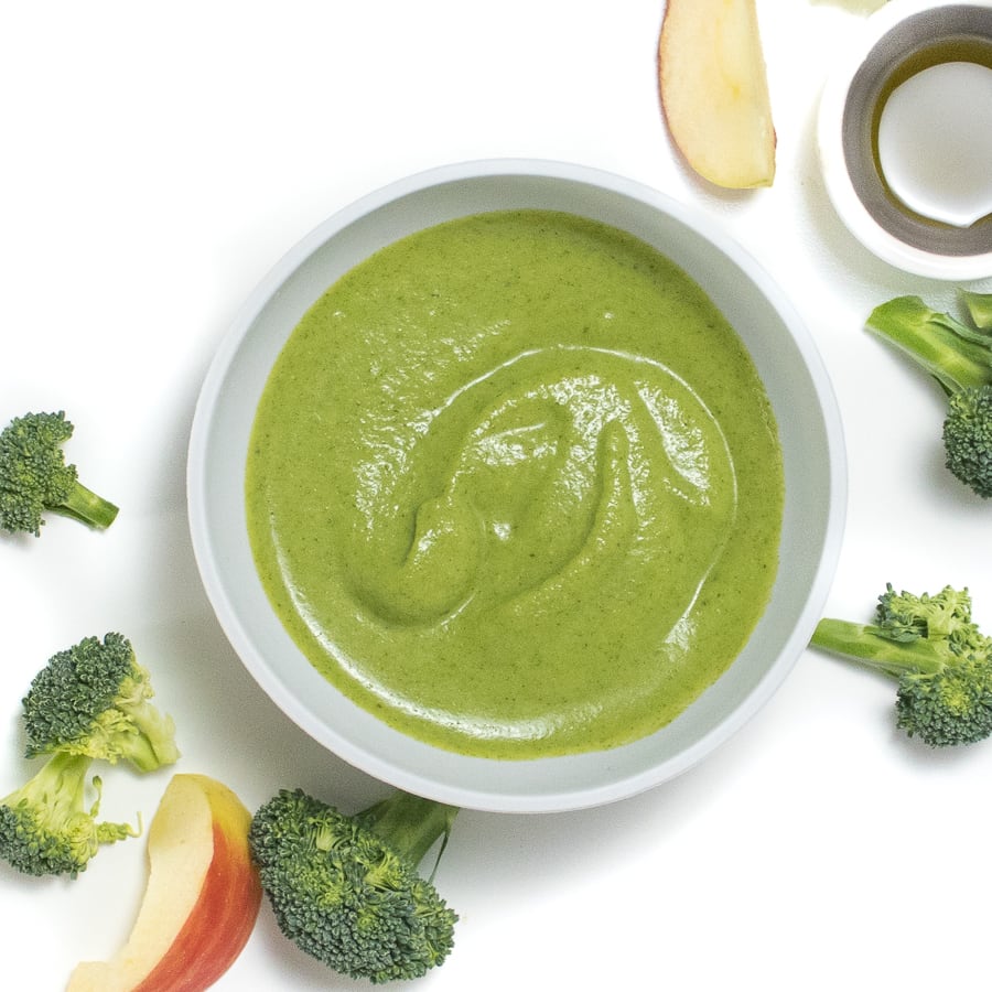 Gray baby bowl with a creamy and smooth broccoli puree in it with broccoli and apples scattered on a white background.