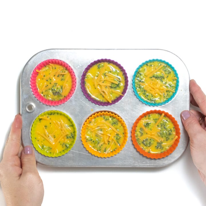 Hands holding a 6-cup muffin tin filled with colorful liners and broccoli egg cups.