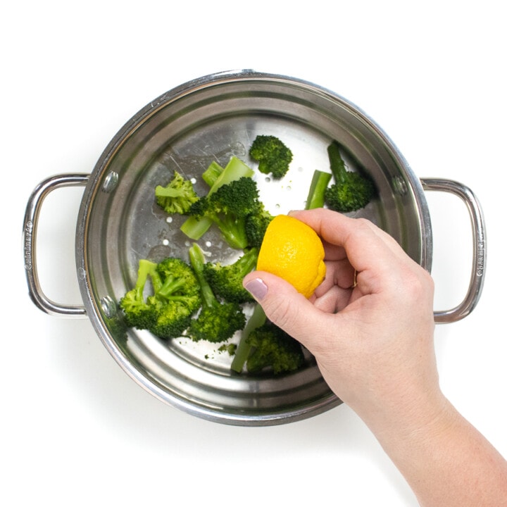 Steamer filled with steamed broccoli with a hand squeezing lemon on to them.