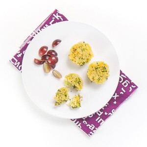 white plate sitting on top of a purple napkin on a white surface. On the plate is 3 mini cheesy broccoli quinoa bites and a small handful of sliced grapes. One of the quinoa bites is halfway eaten with a fork.