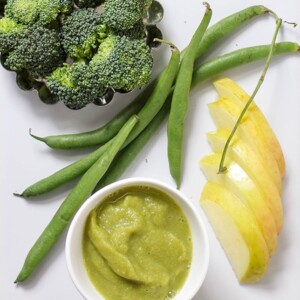 A white cutting board with a spread of produce and small bowl of baby food puree.