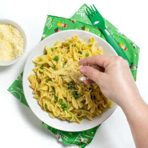 A white bowl of cooked egg noodles and peas with butter and Parmesan. The white bowl is on top of a green kids napkin with the green kids fork with a small bowl of Parmesan next to it. There is a hand sprinkling Parmesan on top.