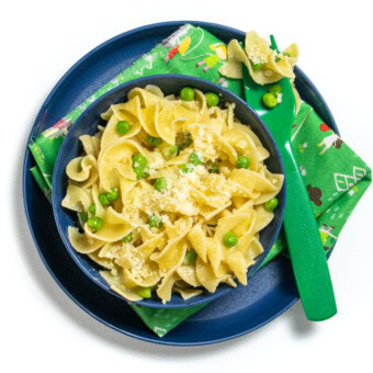 Blue kids plate and bowl with the green fork. The bowl is full with butter noodles with peas and Parmesan and there are noodles on the fork.