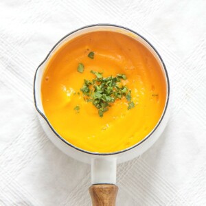 small white saucepan sitting on top of a white tablecloth, inside is a thick butternut squash baby puree with a sprinkle of chopped cilantro on top.