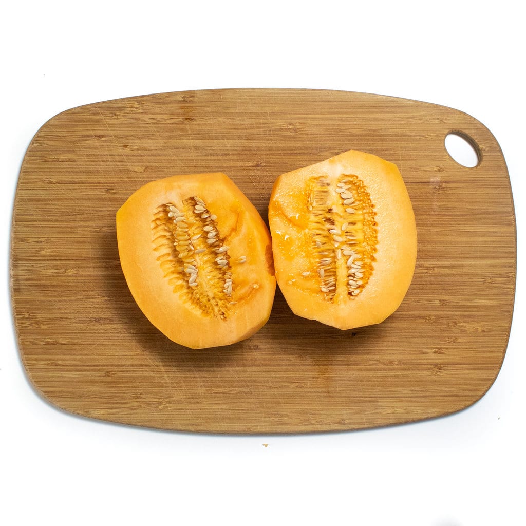 A peeled cantaloupe cut in half with the seeds exposed on a wooden cutting board against a white background.