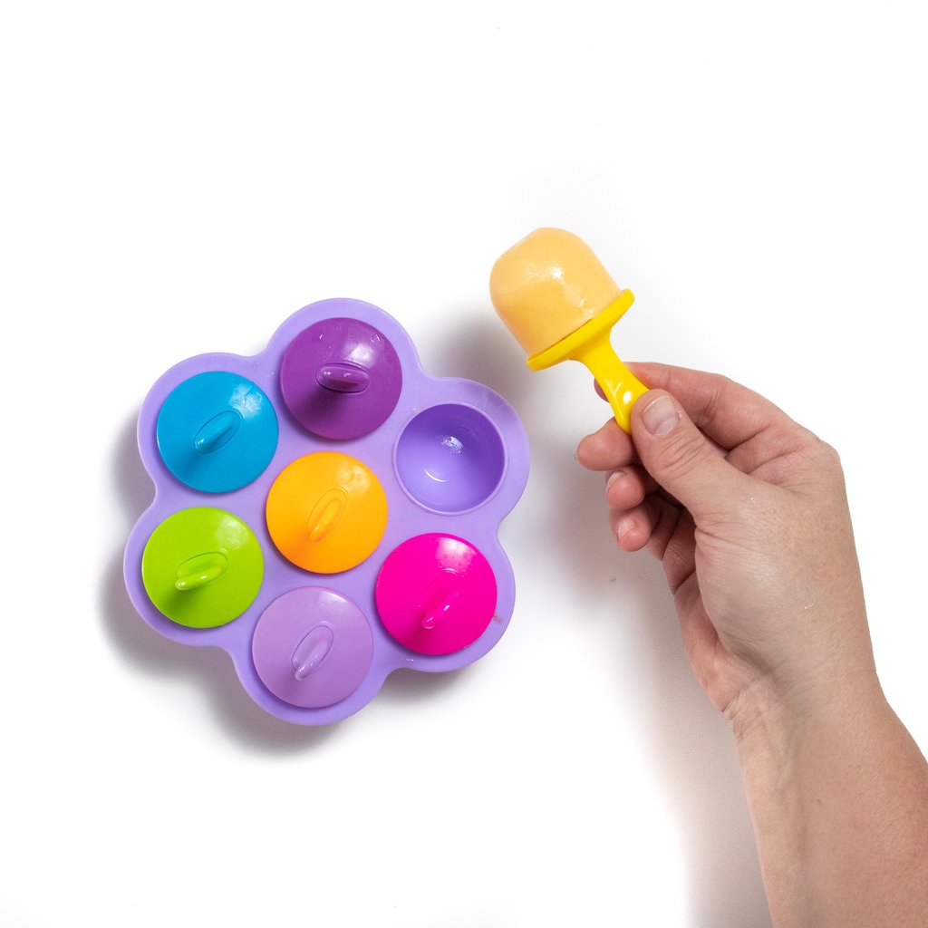 A purple popsicle tray with colorful popsicle sticks and a hand holding a yellow cantaloupe popsicle.