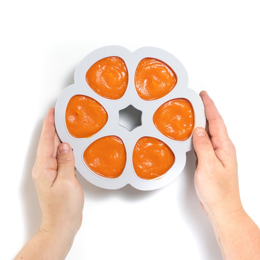 Hands holding a gray baby food storage tray filled with carrot puree.