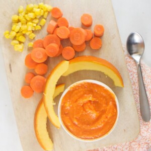 Wood cutting board with a small bowl filled with pumpkin baby food puree with chopped up veggies around it.