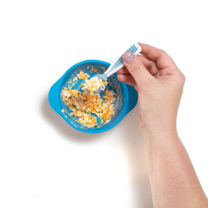 A blue baby bowl filled with mashed carrots, cottage cheese and chives with a hand holding a baby spoonful of this mashed meal.