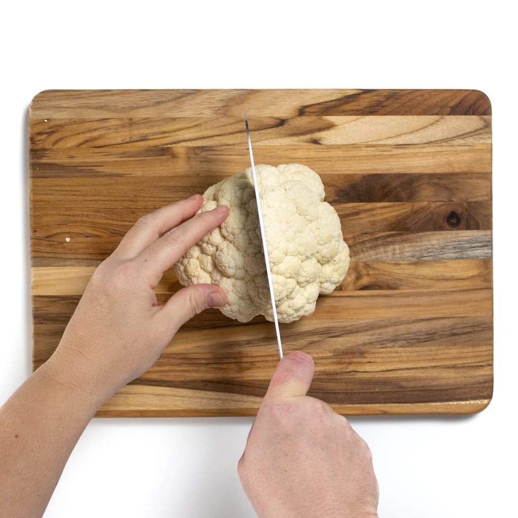 Bored it's a white background two hands are cutting the head of cauliflower in half.