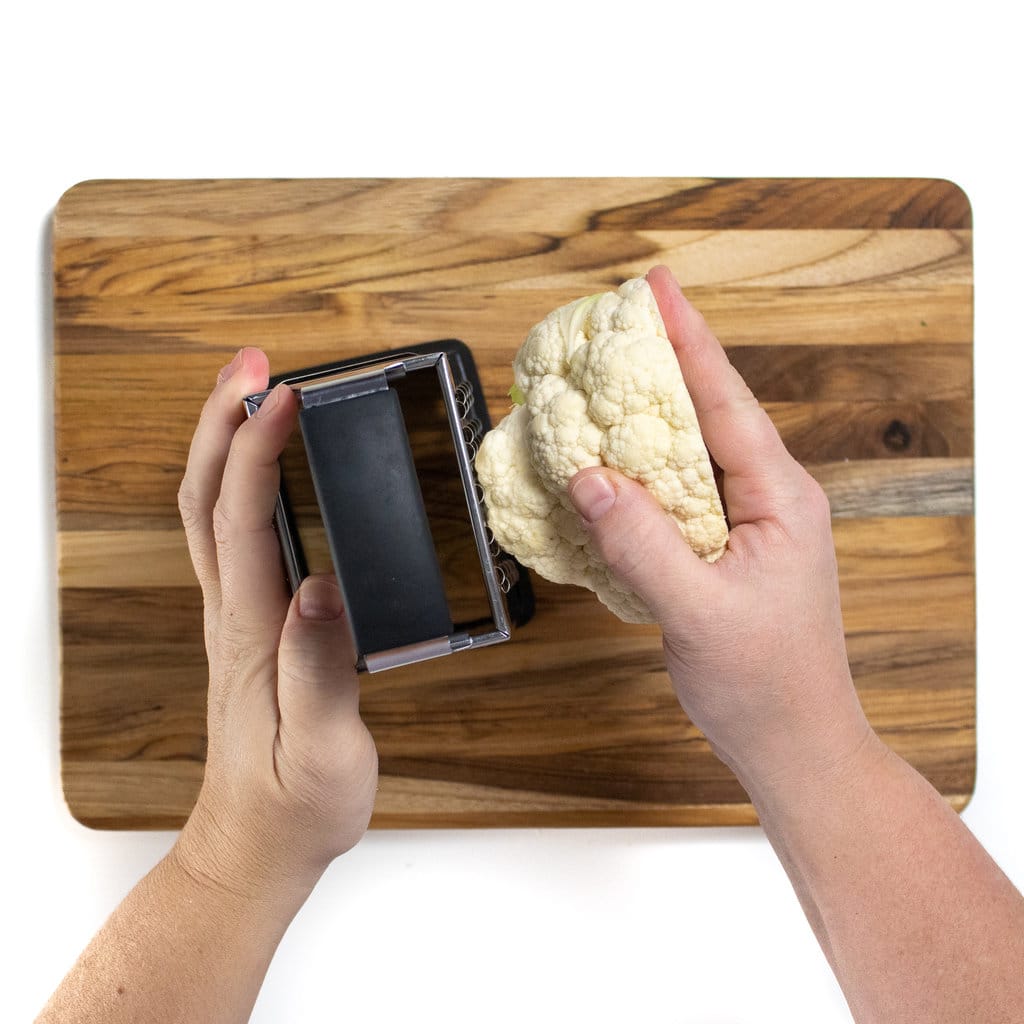 On a wooden cutting board there are two hands grading a cauliflower head to make cauliflower rice.