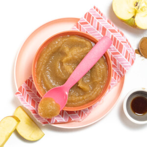 Pink kids bowl on top of a pink napkin filled with homemade applesauce with apples and cinnamon scattered around the bowl.