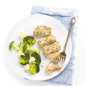 round white plate on top of a blue napkin. On the plate is 4 salmon bites coated in breadcrumbs and herbs. On the side is steamed broccoli and cauliflower.