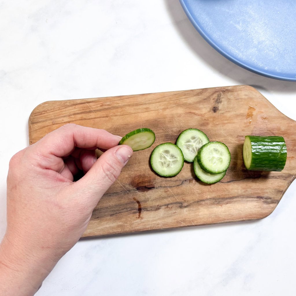 A cutting board on my kitchen counter with thin slices of cucumber for baby, and I'm holding up one showing off the thinness.