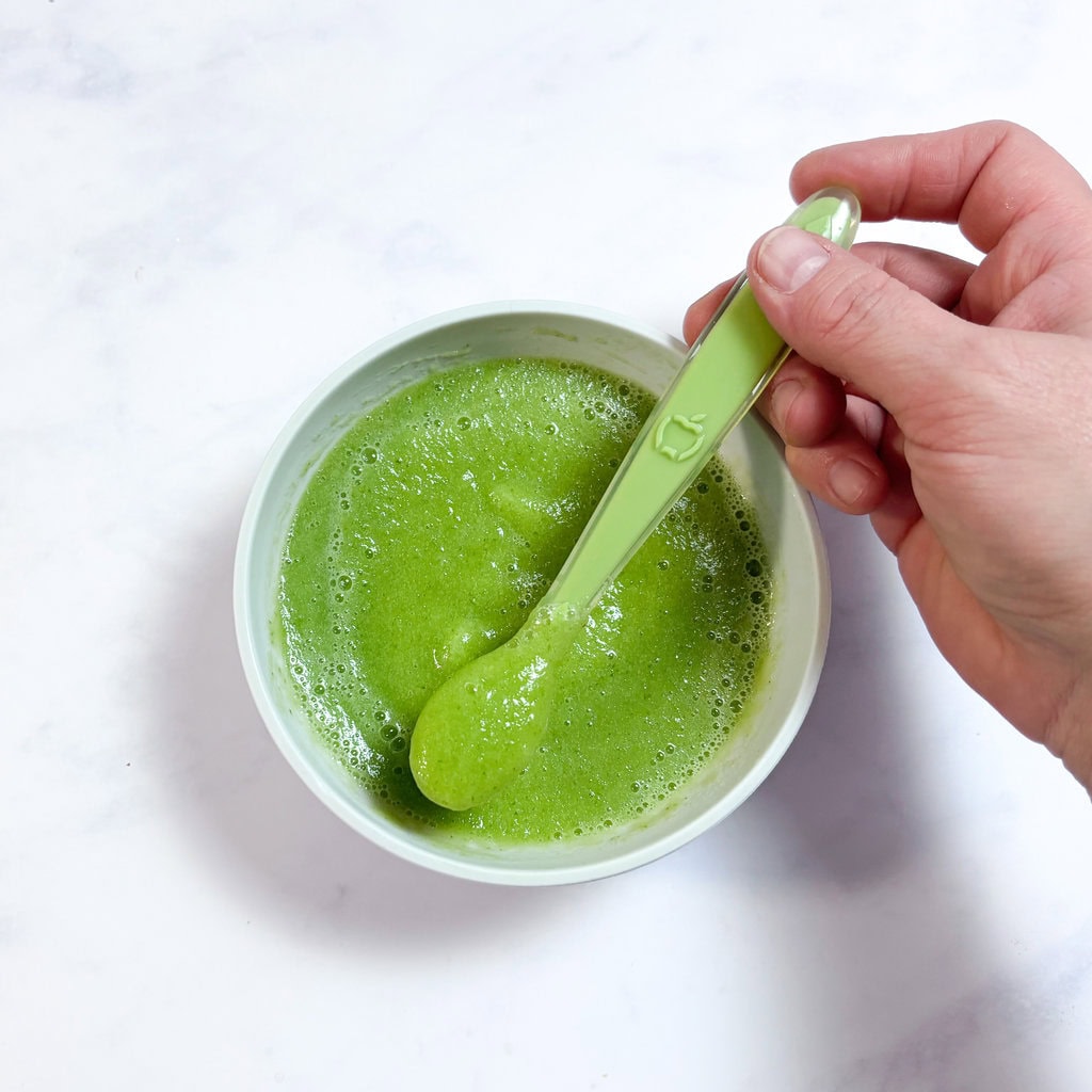 A gray bowl full of cucumber and pear purée for baby with my hand, reaching to scoop up some purée and a green baby spoon.