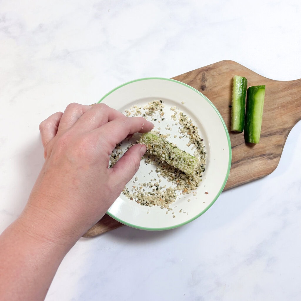 A wooden cutting board with a plate with hemp seeds on it, and I am rolling a thick stick shaped cucumber in the hemp seeds.