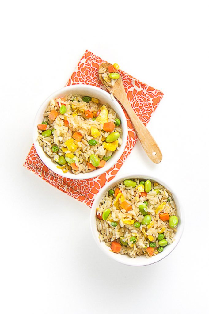 2 white bowls filled with brown rice, vegetables and scrambled egg chunks. Bowls are sitting on a orange flower napkin. There is a wooden spoon on top of one of the bowls with the rice in in. 